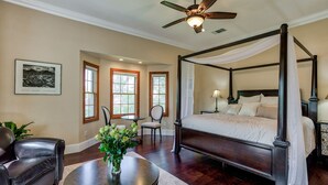 A four-poster canopy bed with a nightstand, a round table with a vase of flowers, a leather sofa, and a ceiling fan in a bedroom with large windows and a framed picture on the wall. - Hope Inn Redding (Redding)