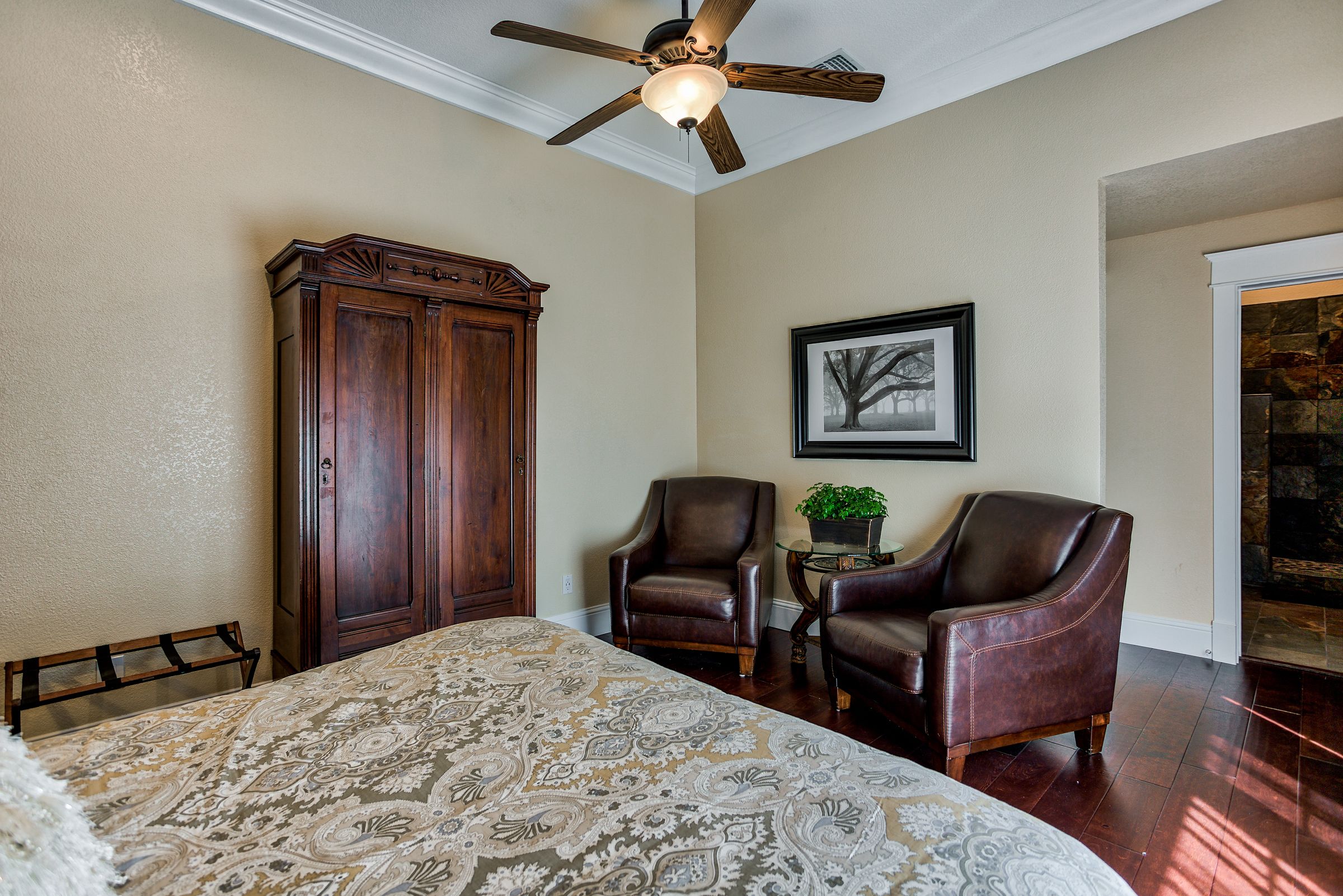 A bedroom with a large bed, two leather chairs, a wooden wardrobe, a ceiling fan, and a framed picture on the wall.
