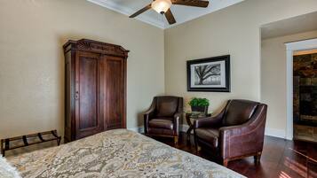 A bedroom with a large bed, two leather chairs, a wooden wardrobe, a ceiling fan, and a framed picture on the wall.