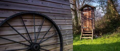 Une structure en bois, avec une grande roue fixée sur le côté, une échelle en bois menant à un petit bâtiment en bois, et une zone herbeuse avec des arbres en arrière-plan.