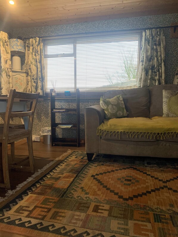 A cozy living room with a patterned rug, a wooden chair, a sofa with cushions, and a window with blinds. - Ceridwen Glamping, Double decker Bus and Yurts (Llandysul)