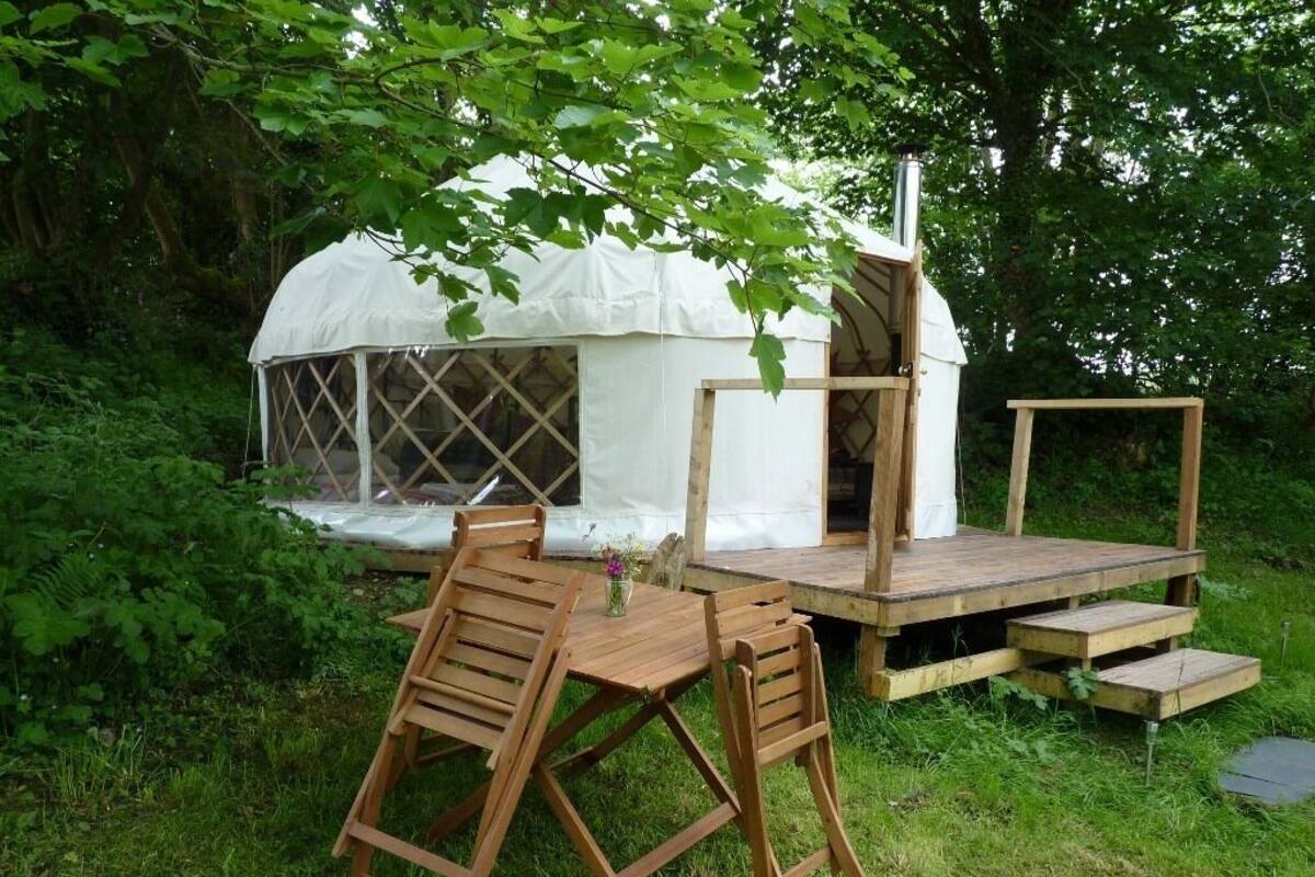 A white tent with a wooden deck and a chimney, surrounded by greenery.