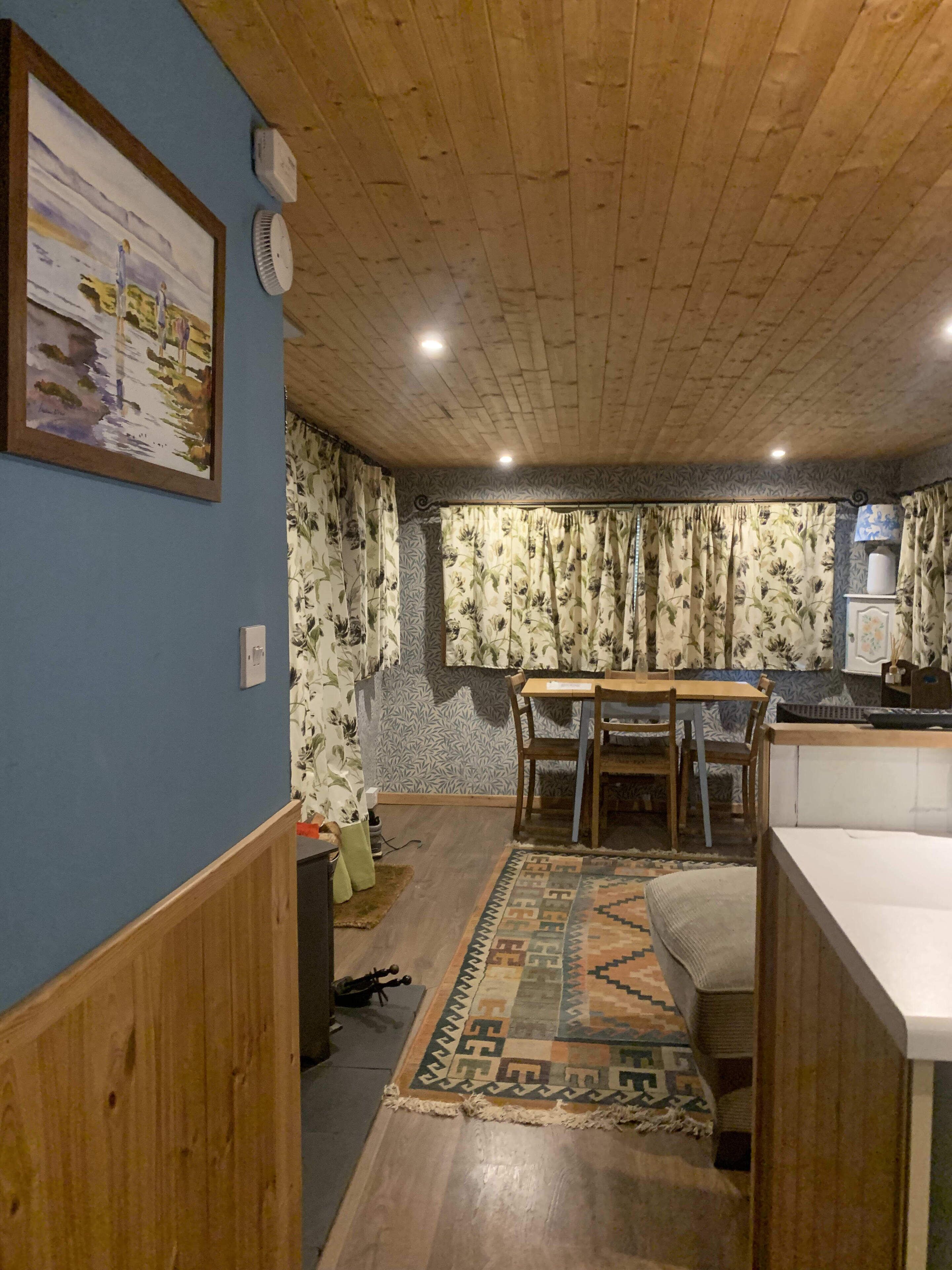 Une pièce chaleureuse avec un plafond en bois, une table à manger avec des chaises, un tapis à motifs et un coin cuisine équipé d’un évier et de placards.