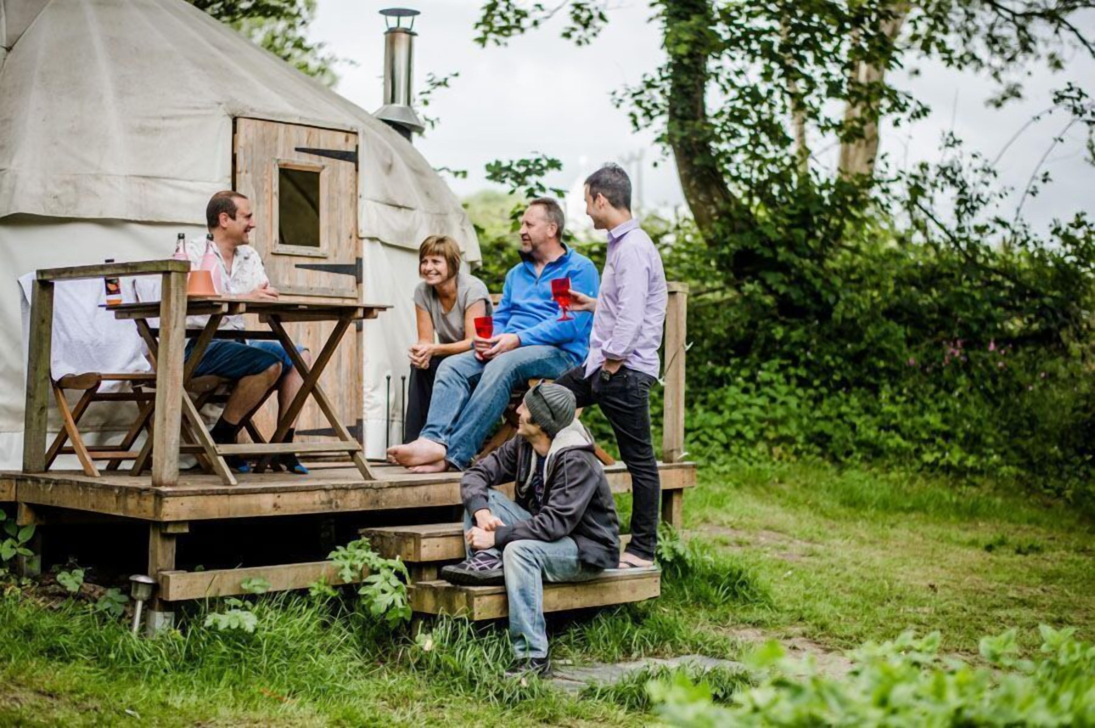 A group of people sitting and standing around a wooden deck attached to a tent-like structure in a grassy area.