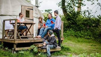 A group of people sitting and standing around a wooden deck attached to a tent-like structure in a grassy area.