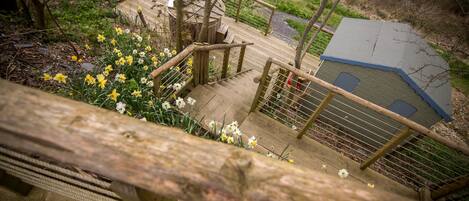 A wooden staircase leading to a small wooden structure with a metal roof, surrounded by a garden with yellow and white flowers.