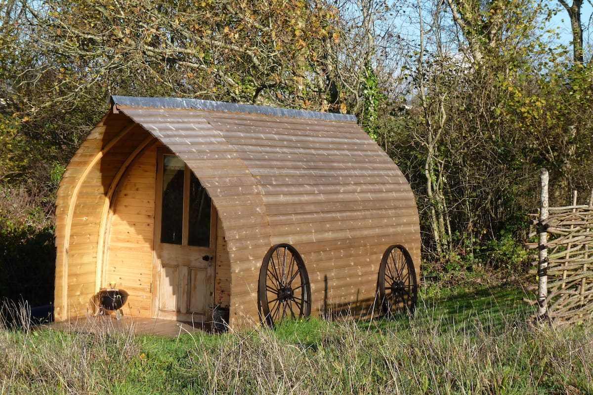 A wooden cabin with a curved roof and large wheels on the sides, surrounded by trees and grass.