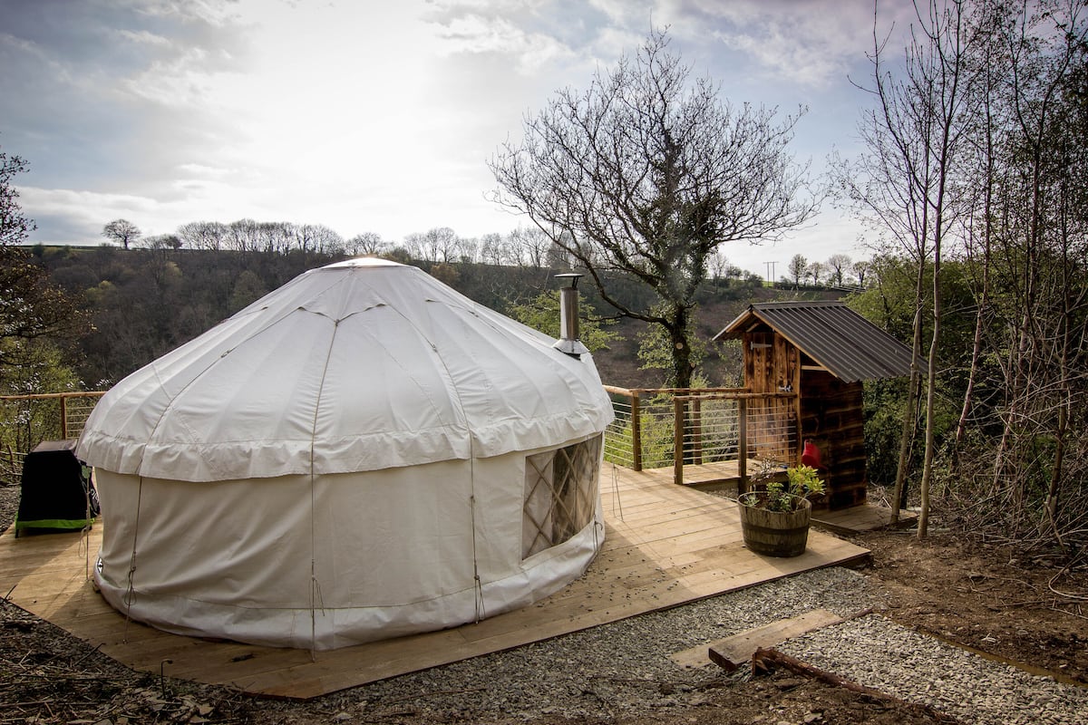 A white yurt with a wooden deck, a small wooden cabin, and a view of the surrounding hills.