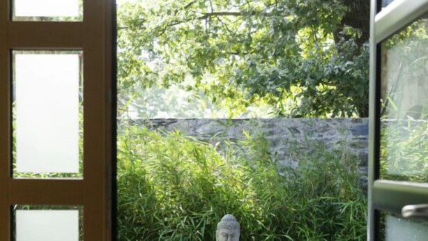 A wooden deck with a stone Buddha statue, surrounded by lush greenery and a stone wall. - La Balade des Marais (Saint-Molf)