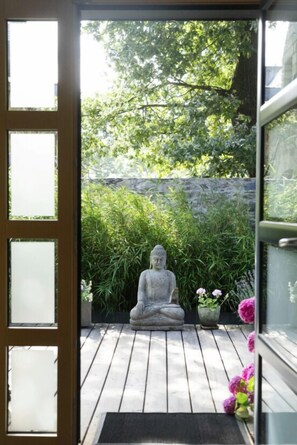 Una terraza de madera con una estatua de Buda de piedra, rodeada de exuberante vegetación y un muro de piedra.