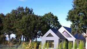 A modern house with a sloped roof and large windows, surrounded by a well-maintained lawn and tall trees. - La Balade des Marais (Saint-Molf)