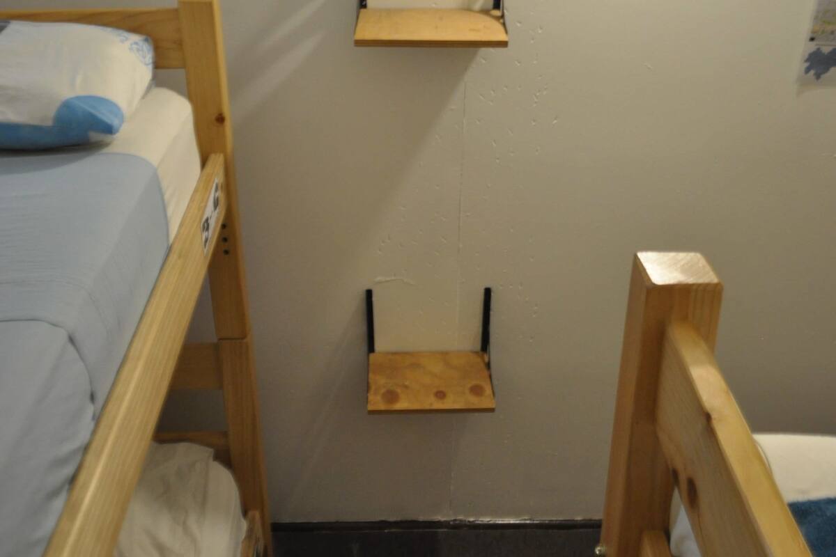 Una habitación pequeña y estrecha con dos literas, una estantería de madera y una lámpara fijada a la pared.