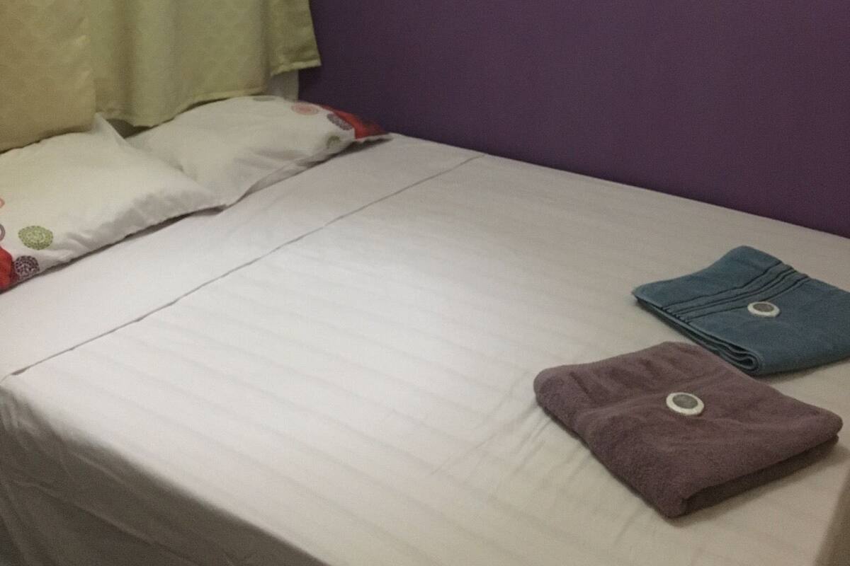 Una cama individual con colchón blanco, pared morada y cortina amarilla.