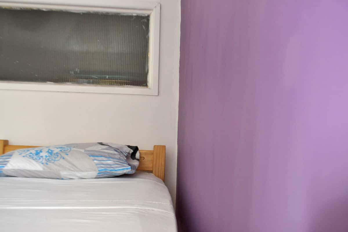Una cama con cabecera de madera, una ventana con marco blanco y una pared morada.