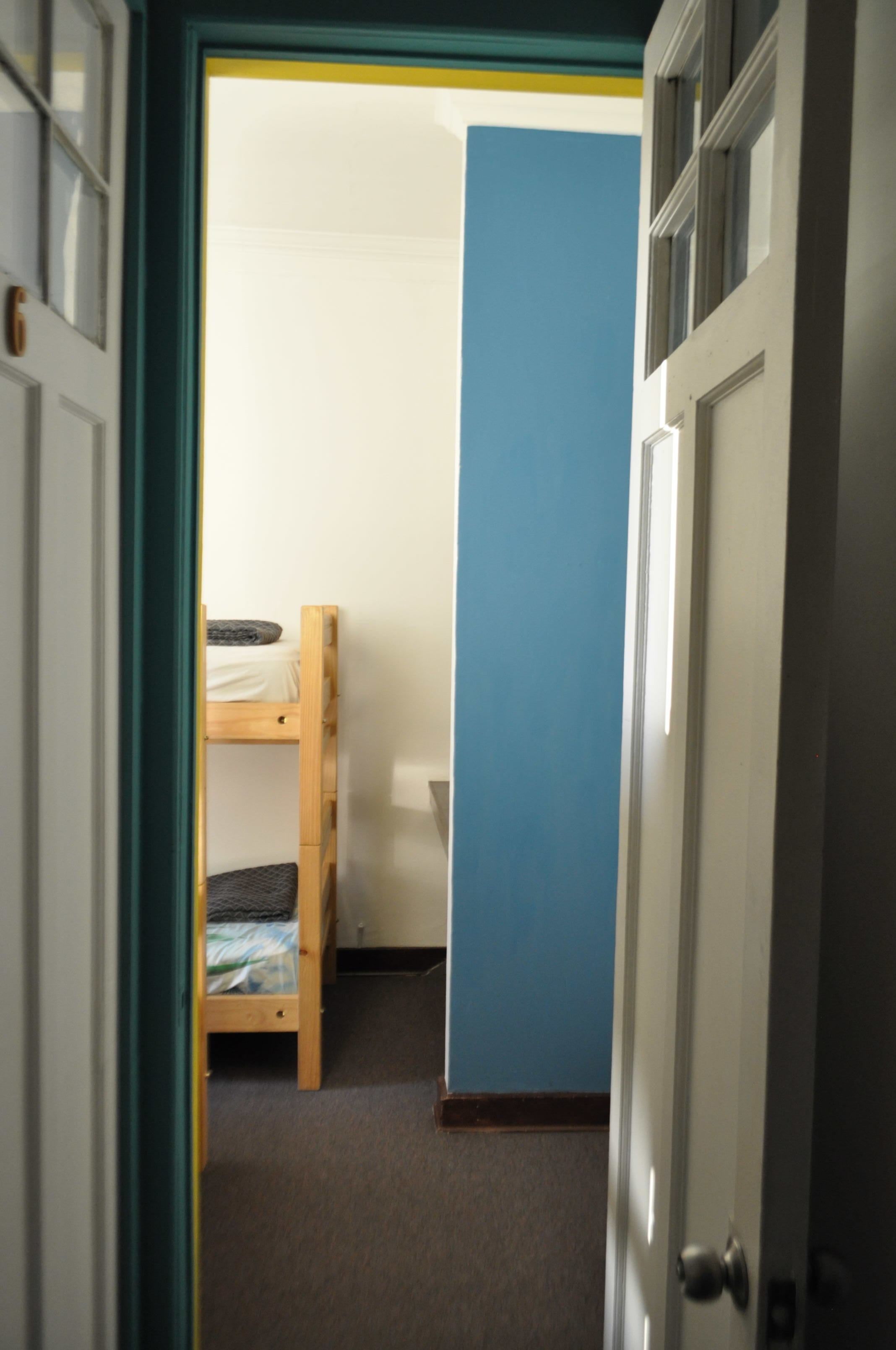 Un pasillo estrecho con una cama litera al fondo, una pared azul y una puerta cerrada.