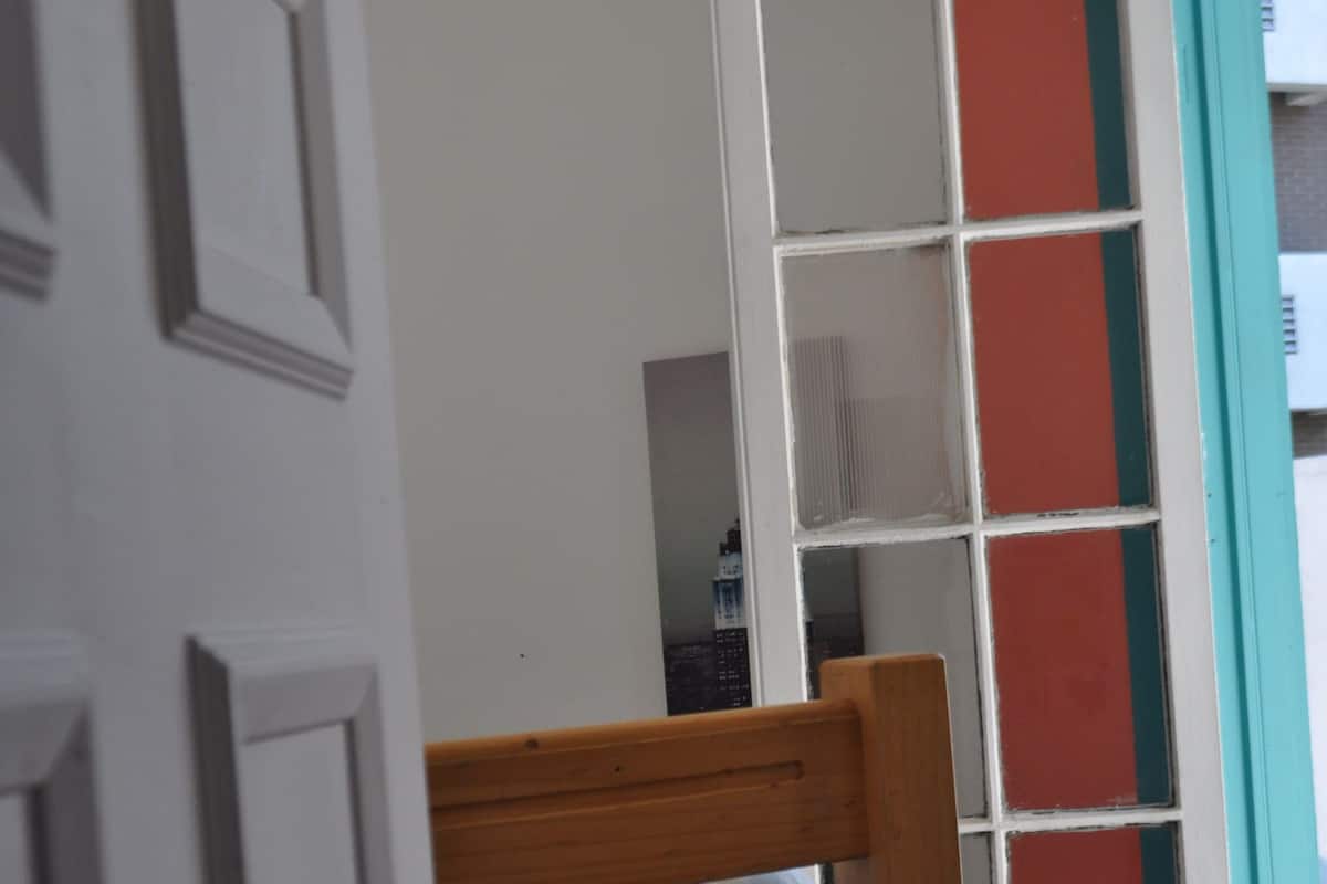 Una habitación con una cama nido de madera, una pequeña silla de madera, una puerta blanca y un ventanal con marco rojo.