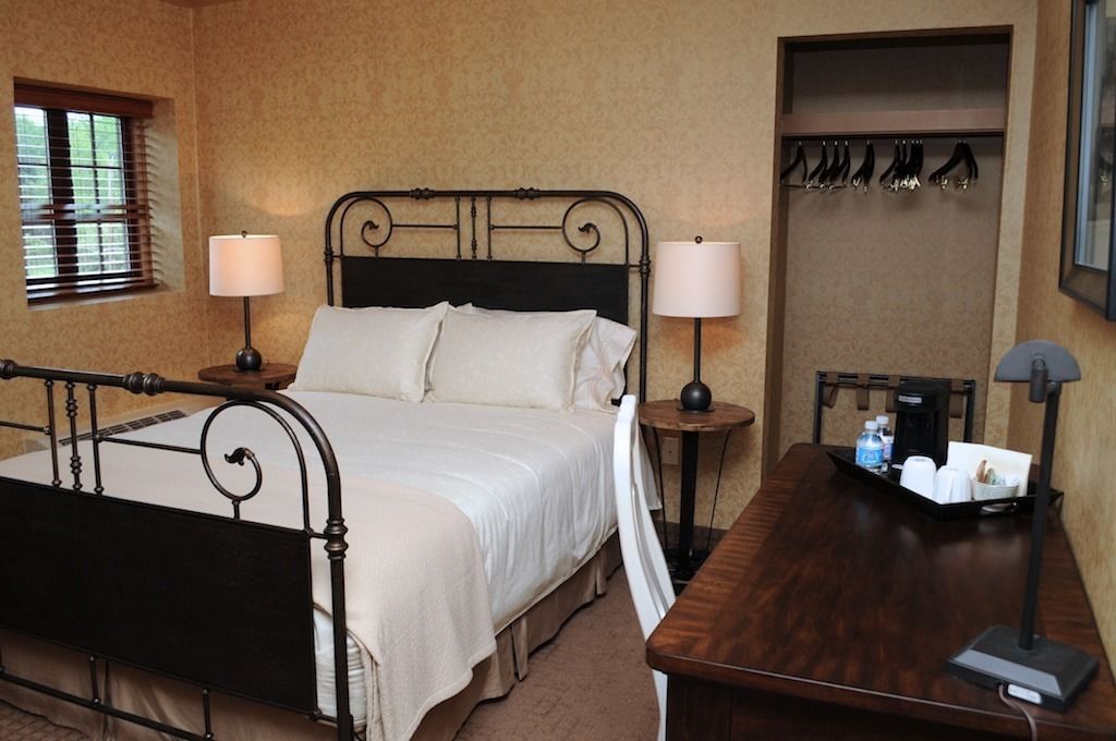 A neatly arranged hotel room with a bed, two bedside tables with lamps, a wooden side table, a coat rack, and a small table with toiletries.