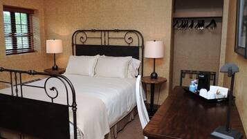 A neatly arranged hotel room with a bed, two bedside tables with lamps, a wooden side table, a coat rack, and a small table with toiletries.