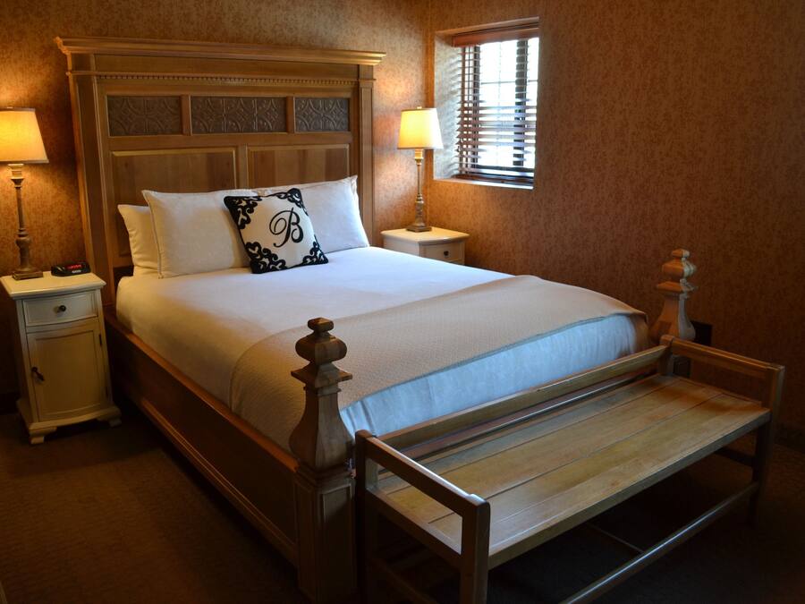 A neatly made bed with a wooden headboard, two bedside tables with lamps, and a window with blinds.
