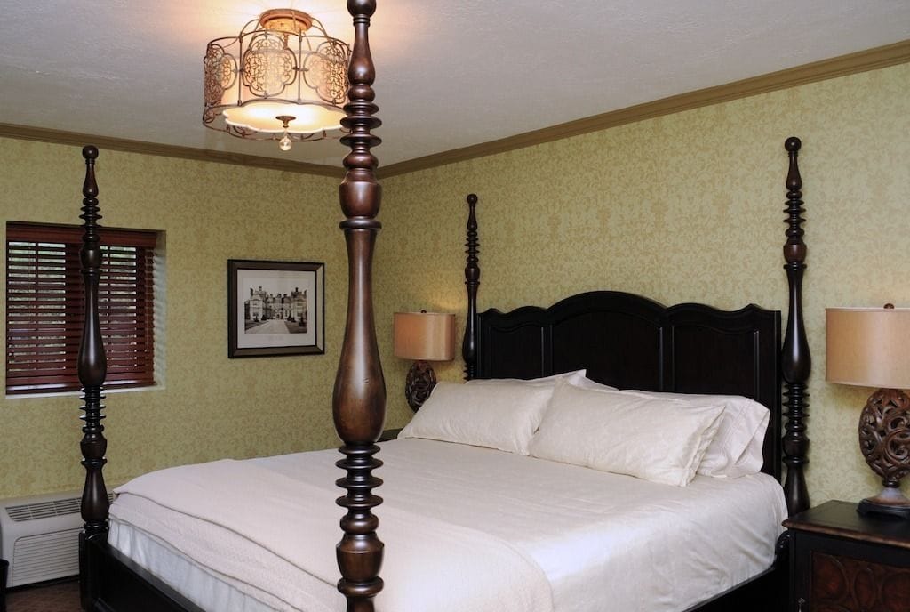 Een hemelbed met een donker houten frame en een witte spreidekker. Aan de linkerzijde van het bed hangt een ingelijst schilderij.