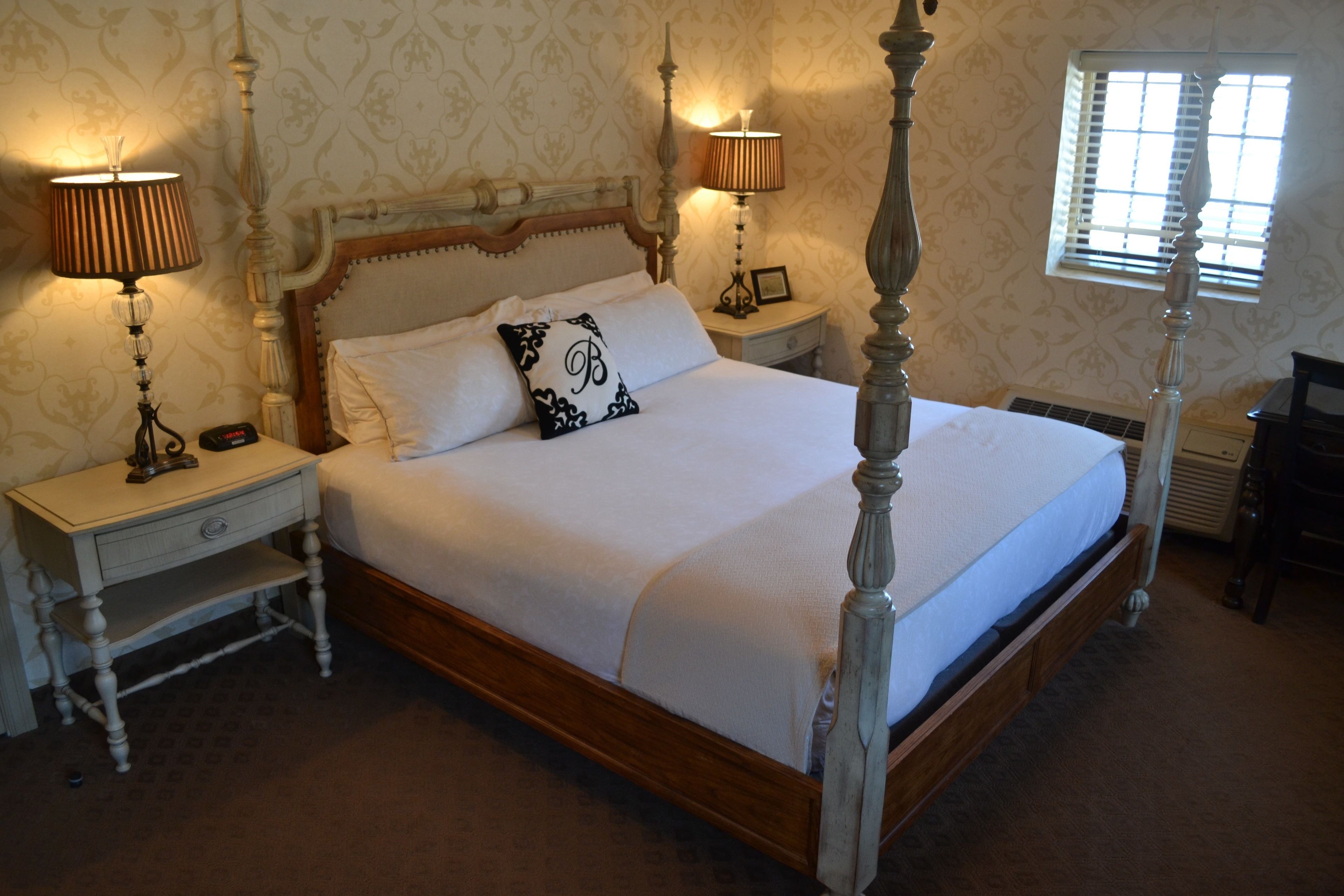 A four-poster bed with a wooden frame and white bedding, flanked by two bedside tables with lamps, in a room with patterned wallpaper and a window with blinds.