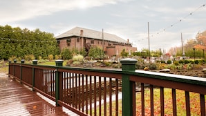 A wooden deck with a green railing, a brick building, and a garden area with trees and plants. - The Beaumont Inn (Dallas)