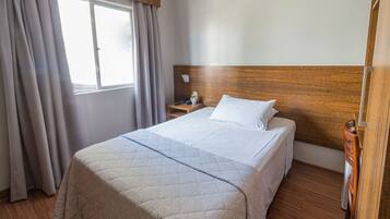 Una cama bien tendida con un cabecero de madera, una mesita de noche con una lámpara y una ventana con cortinas.