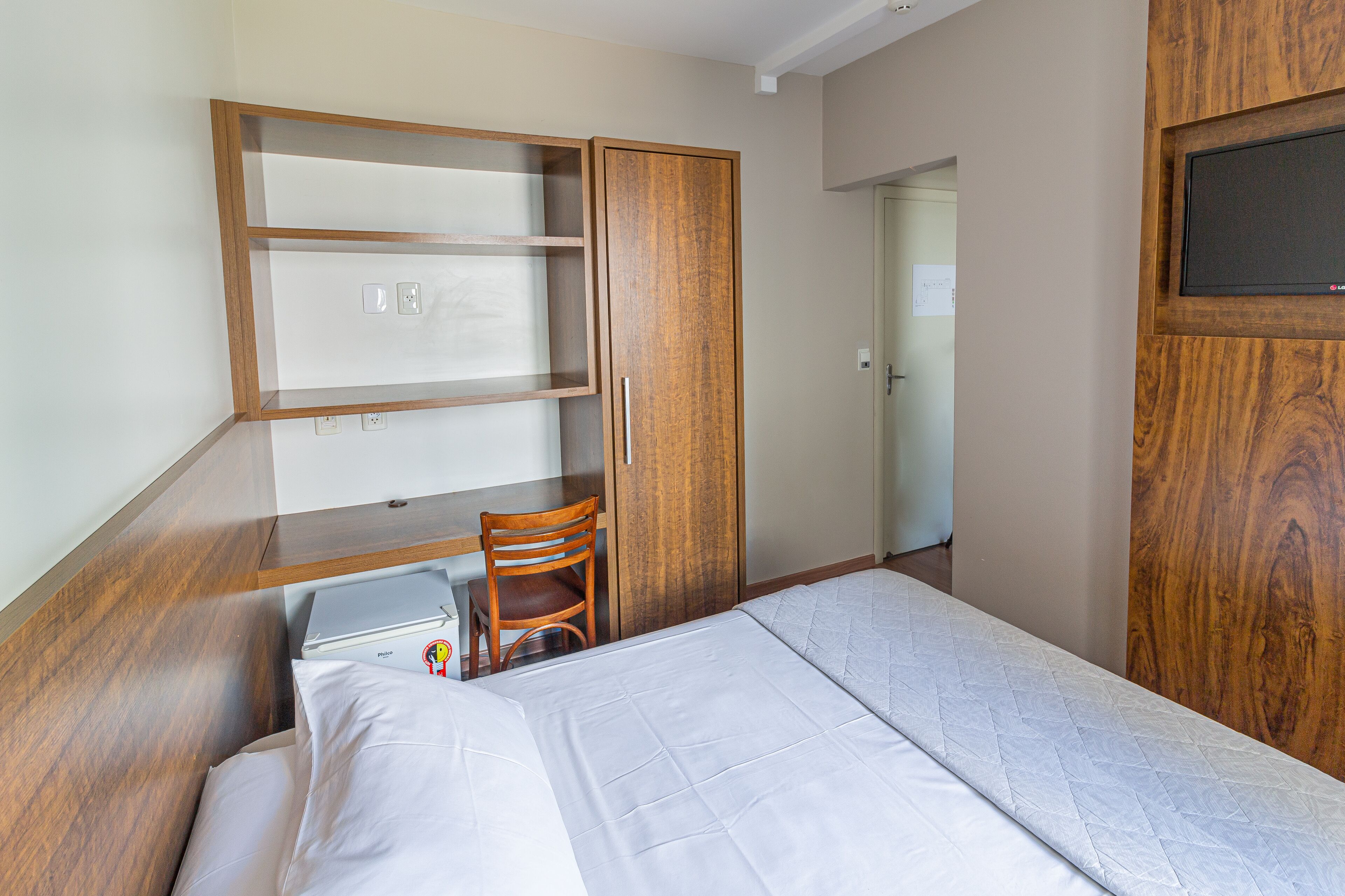 Un dormitorio con una cama, un armario de madera, un escritorio con una silla y un televisor instalado en la pared.