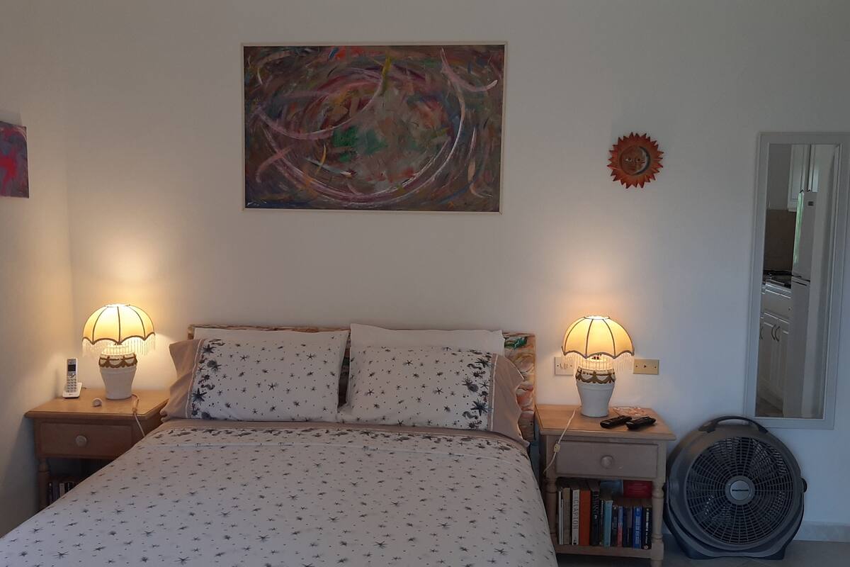 A bedroom with a bed, two bedside lamps, a wall clock, a small table with books, and a wall-mounted fan.
