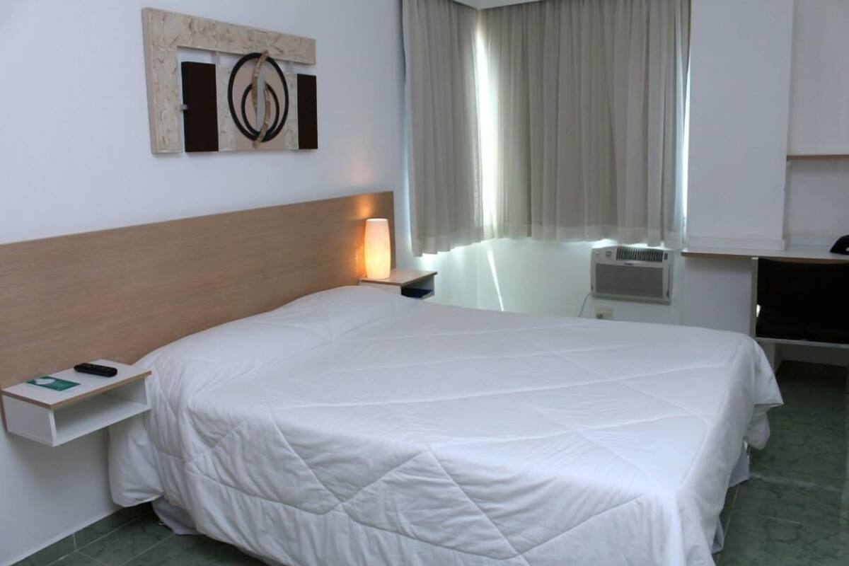 Une chambre d’hôtel avec un lit, une table de chevet avec une lampe, une œuvre d’art accrochée au mur et une fenêtre avec des rideaux.