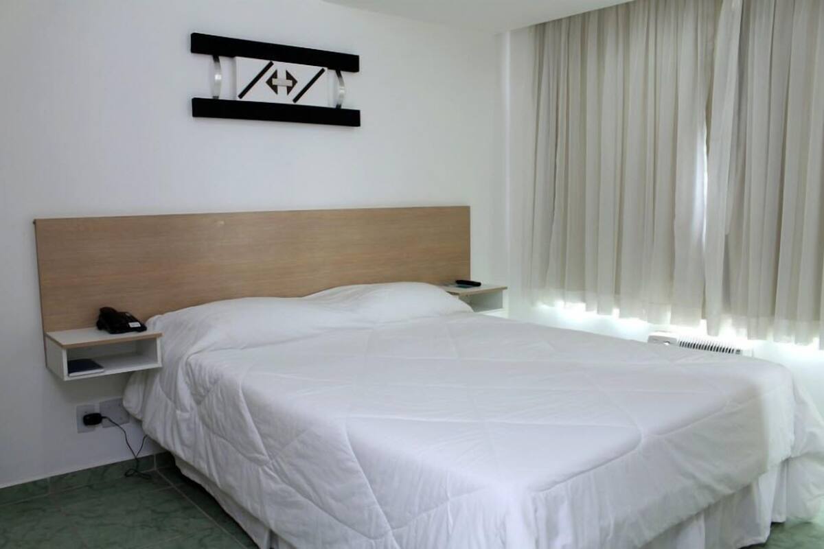 Une chambre d’hôtel avec une tête de lit en bois, un couvre-lit blanc, une table de chevet avec un téléphone et une fenêtre ornée de rideaux légers.