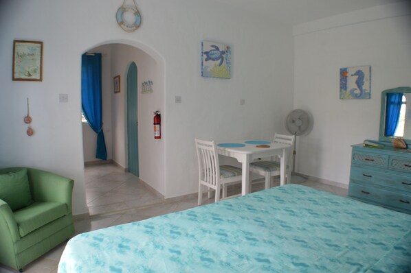A room with a bed, a green armchair, a dining table with chairs, a dresser, and a fan. - Two Palms Apartment (Silver Sands)
