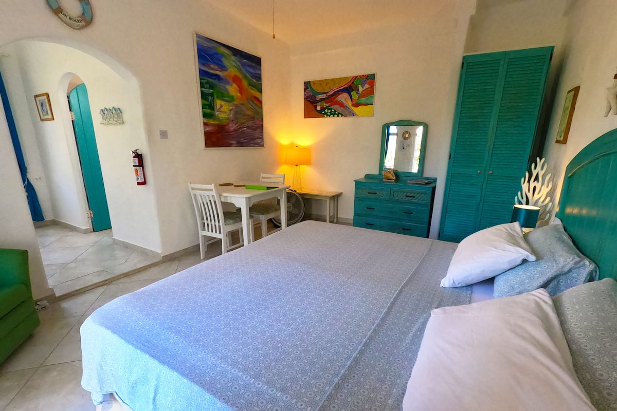 Quarto com cama, escrivaninha, cadeira, abajur, espelho e porta.