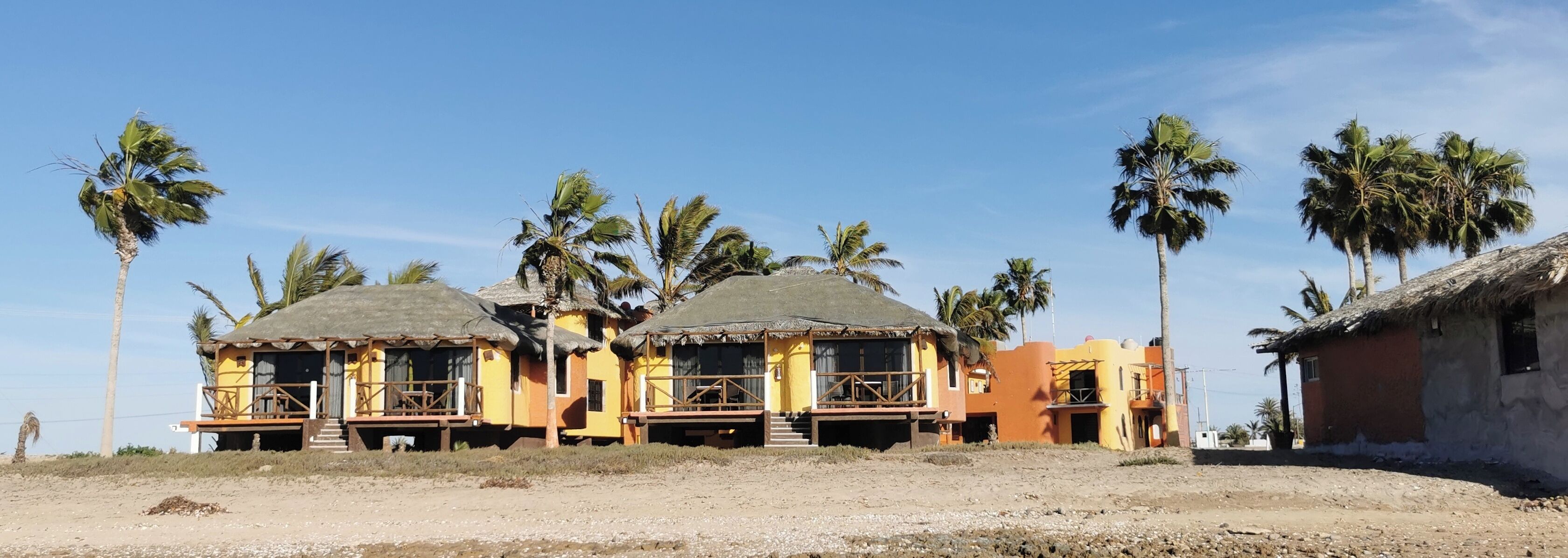 Una propiedad frente a la playa con estructuras de techo de paja, palmeras y un primer plano arenoso.