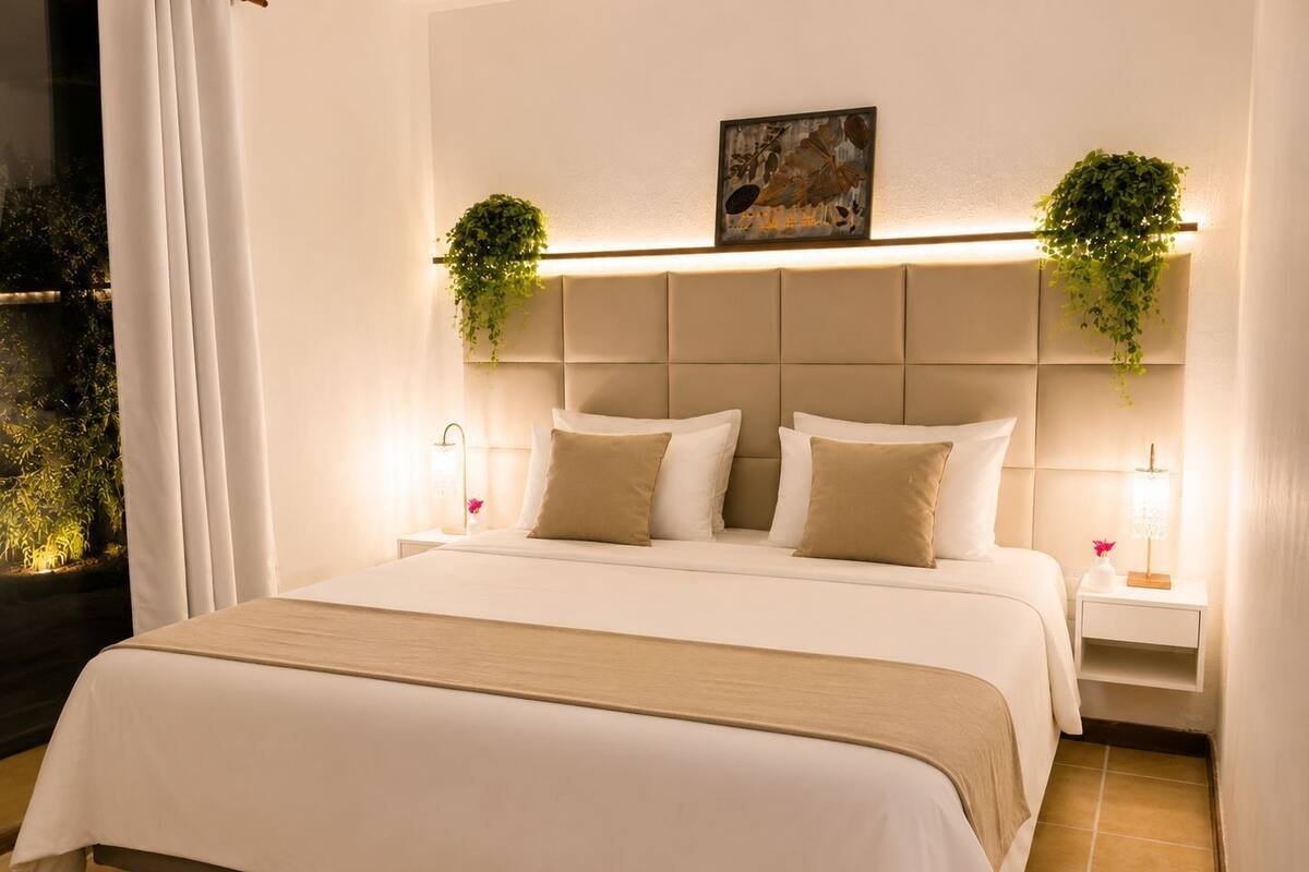 Une chambre d’hôtel avec un grand lit, deux tables de chevet, une tête de lit agrémentée d’une plante et une fenêtre avec des rideaux.
