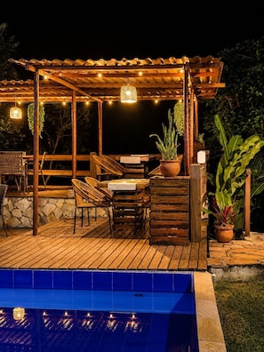 Um deck de madeira com móveis para área externa, uma pérgula, luzes de fada e uma piscina.