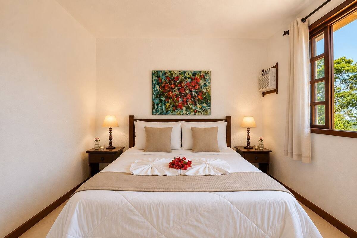 Un lit bien fait, agrémenté d’un arrangement floral, deux lampes de chevet, un tableau au mur et une fenêtre avec des rideaux.