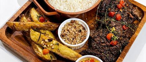 Una bandeja de madera con arroz, patatas asadas, un bistec con tomates cherry, un cuenco de frutos secos y un recipiente con salsa.