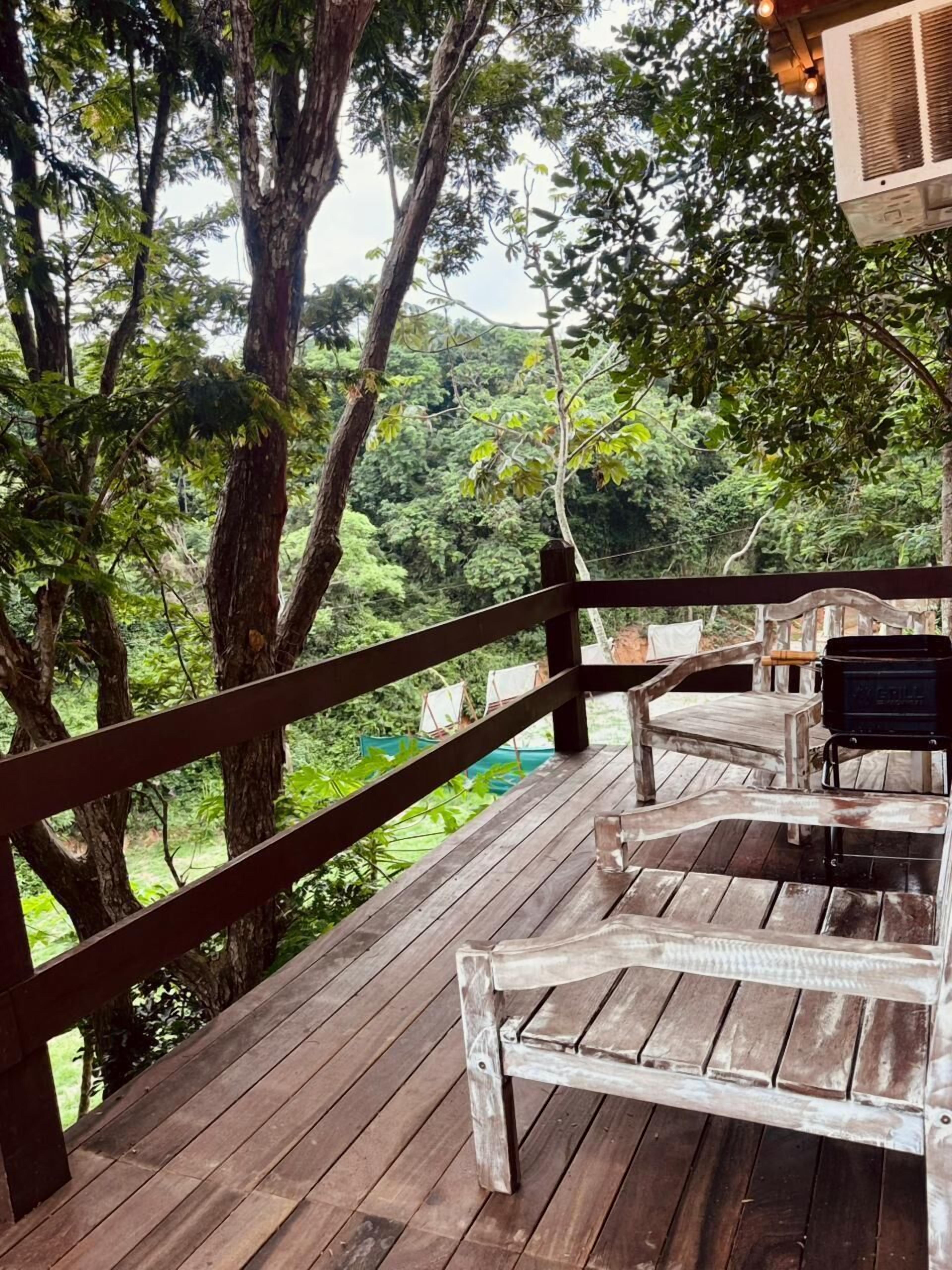 Una terraza de madera con un banco y una parrilla, rodeada de árboles.