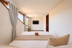 Quarto com cama, televisão, mesinha com cesta e janela com cortinas.