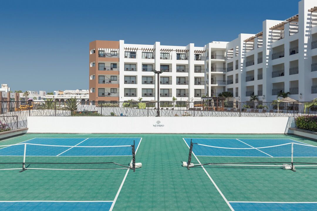Una cancha de tenis con dos mallas, rodeada por un edificio blanco con balcones.
