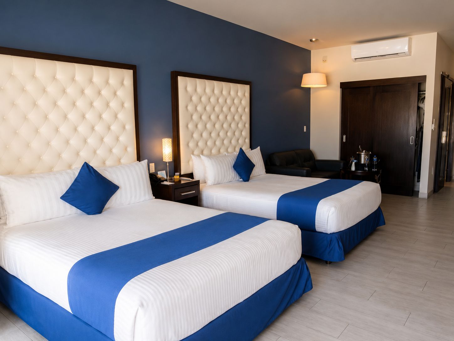 A hotel room with two beds, a dark-colored sofa, a nightstand with a lamp, and a wardrobe.