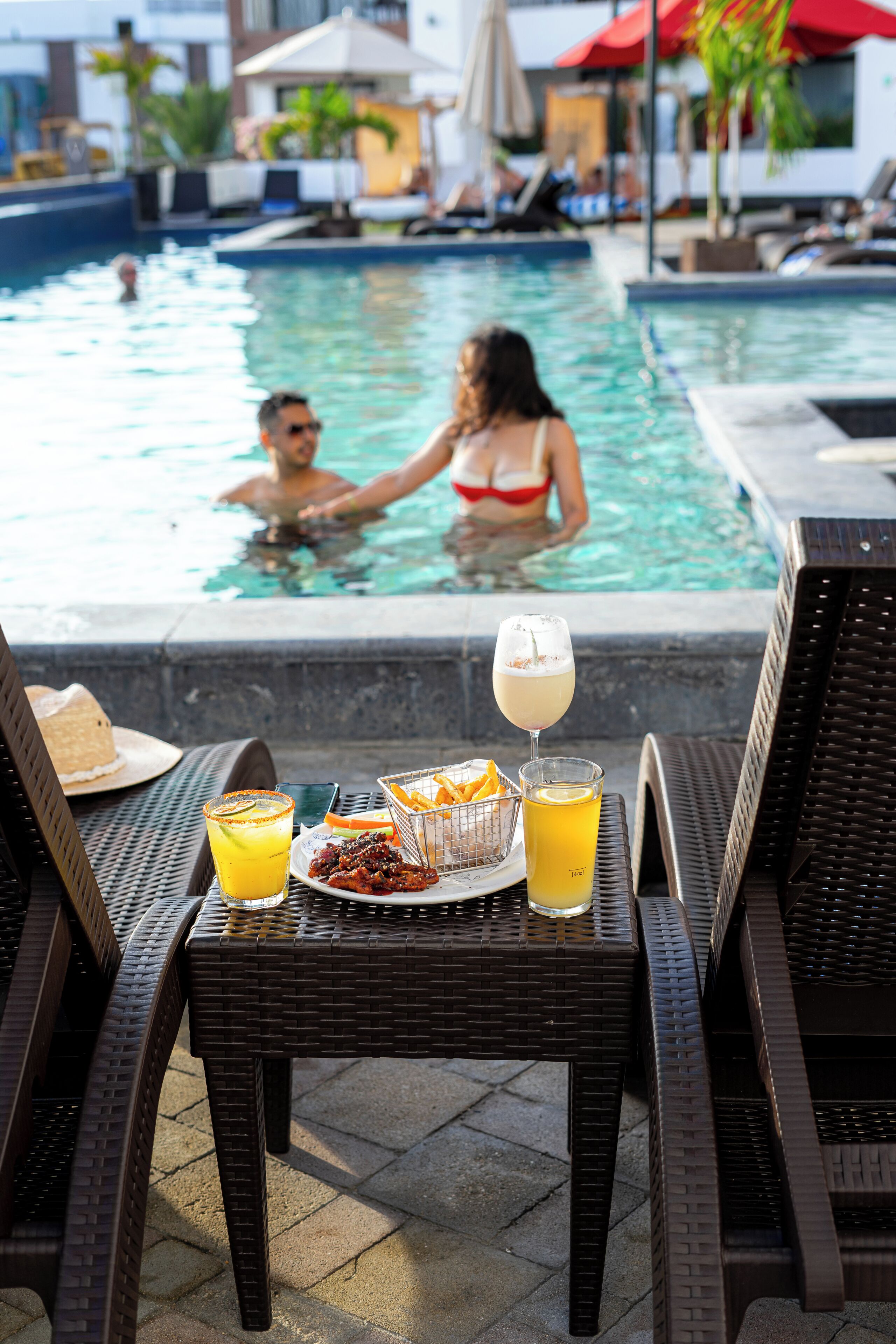 A poolside setting with lounge chairs, a table with a drink and snacks, and a pool in the background.