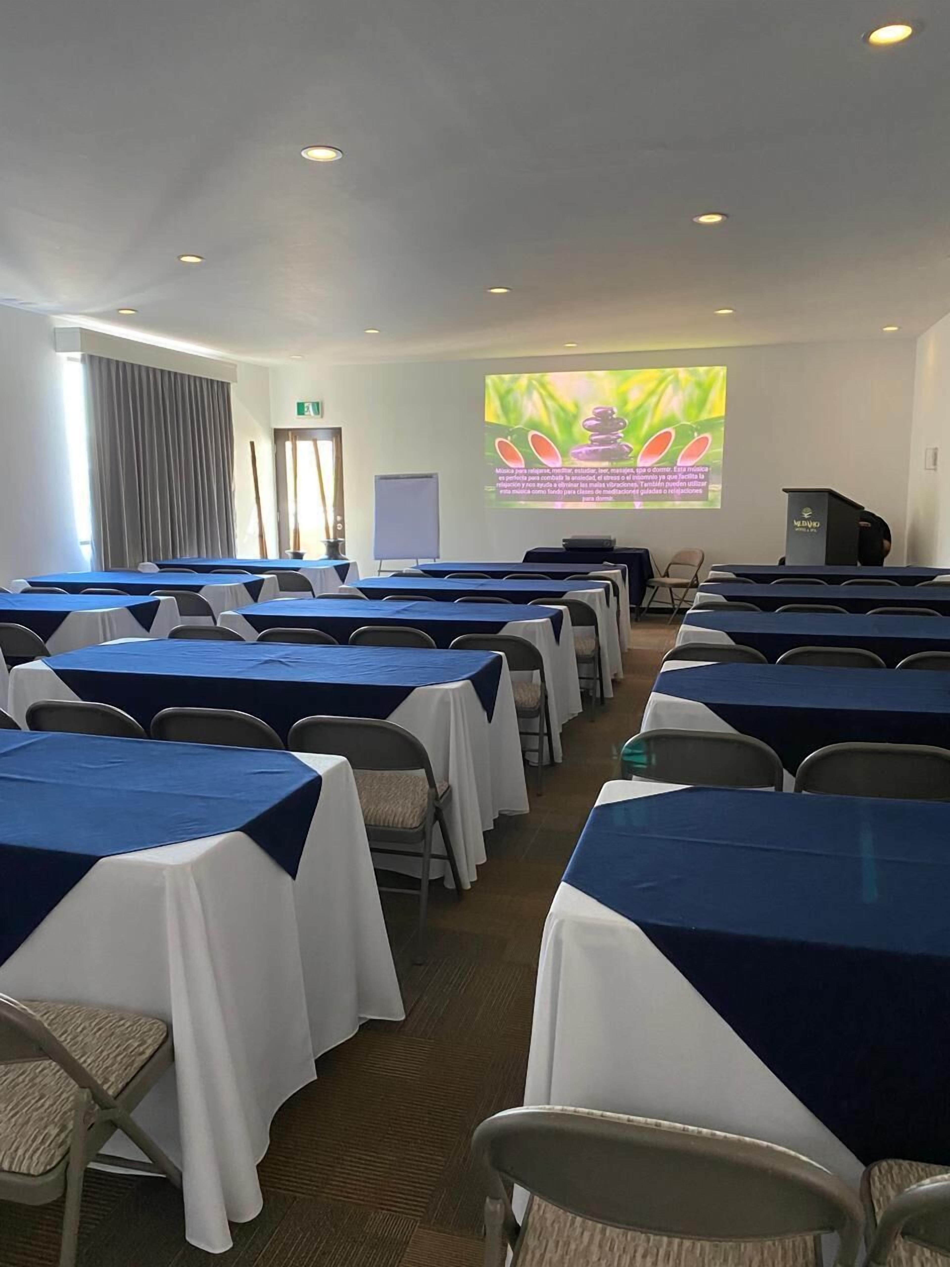 Un salón de conferencias con filas de mesas cubiertas con manteles azules y blancos, equipado con sillas, una pantalla proyectora mostrando una presentación y un atril.