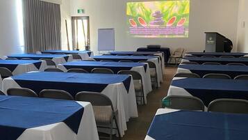 Un salón de conferencias con filas de mesas cubiertas con manteles azules y blancos, equipado con sillas, una pantalla proyectora mostrando una presentación y un atril.