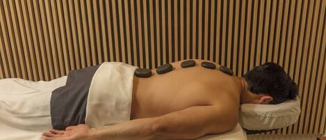 A person receiving a back massage with stones on their back, lying on a bed with a striped wall in the background.