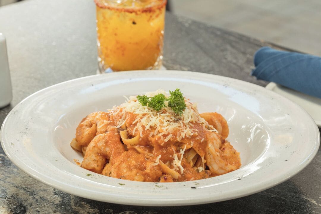 A plate of pasta with a tomato-based sauce, garnished with cheese and parsley, on a table with a glass of a beverage in the background.