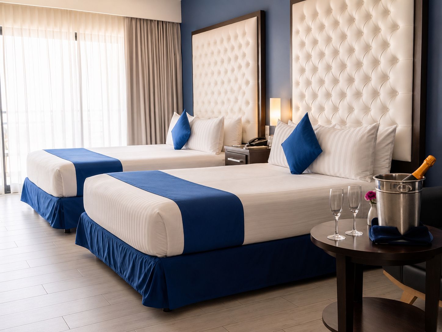 A hotel room with two beds, a minibar, and a round table with glasses and a bottle.
