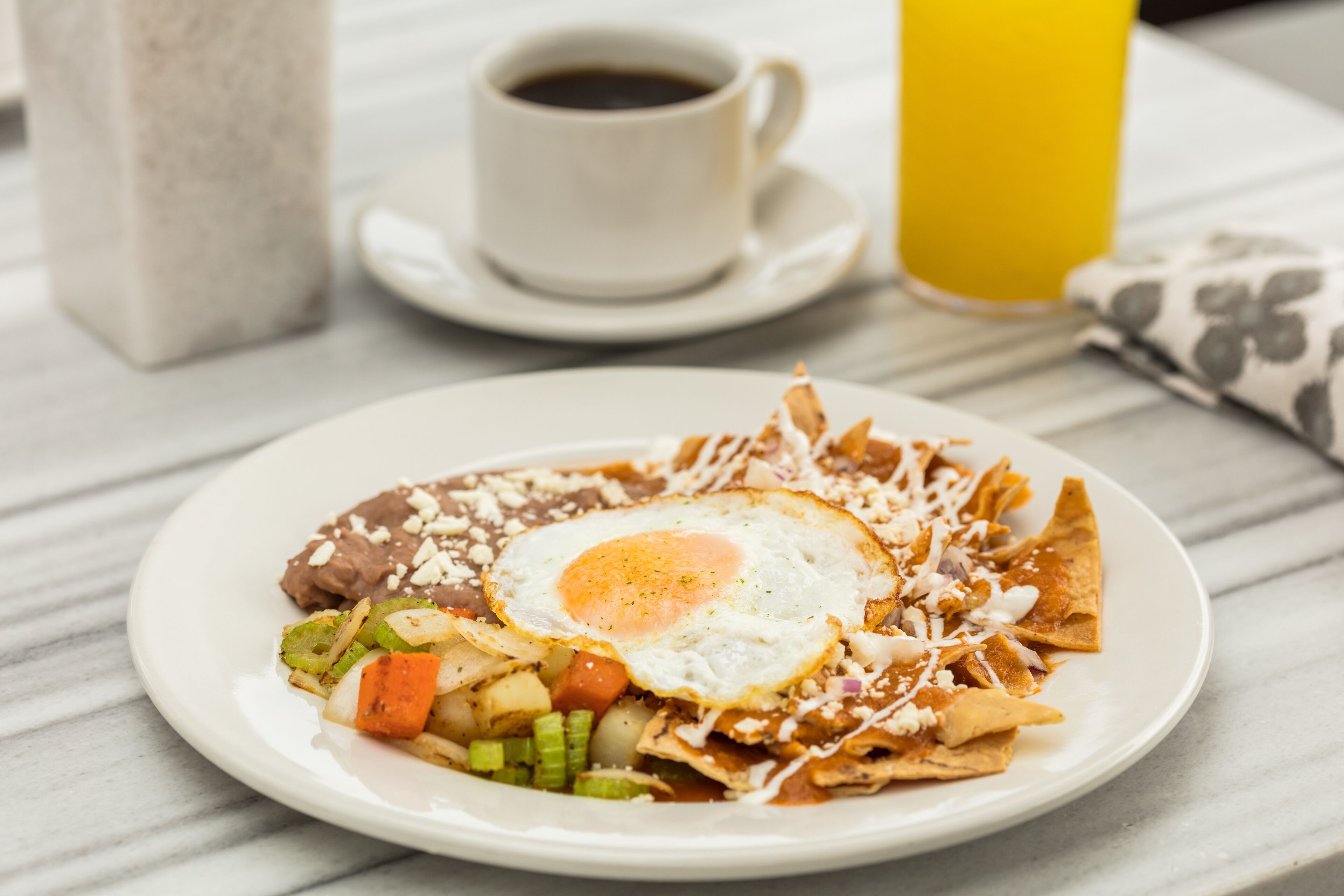 A plate of nachos with a fried egg, beans, and vegetables, accompanied by a cup of coffee and a glass of orange juice on a marble table.