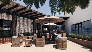 An outdoor seating area with wicker furniture and a large umbrella, surrounded by planters and a building facade. - Medano Hotel and Spa (Cabo San Lucas)